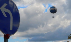 Das Foto zeigt einen großen Luftballon vor einem  Himmel, hier in Berlin. Im Vordergrund ist ein irdisches Verkehrschild, das das Abbiegen nach links vorschreibt.